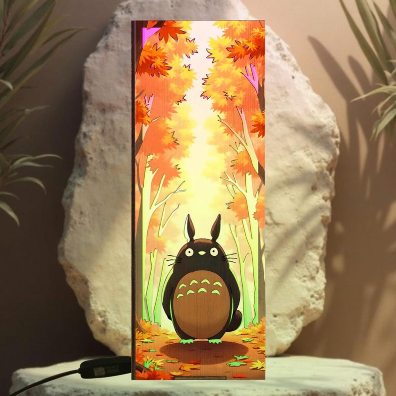 Puede incluir: L&aacute;mpara rectangular de madera con un personaje de dibujos animados de Totoro en una escena de bosque con follaje oto&ntilde;al. La l&aacute;mpara tiene un cable de alimentaci&oacute;n negro y est&aacute; sobre un fondo neutro.