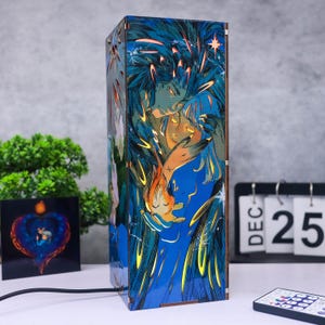 May include: A rectangular wooden lamp with a blue and orange illustration of two figures embracing. The lamp has a warm glow and is plugged in with a black cord. A small square art piece and a calendar are in the background.