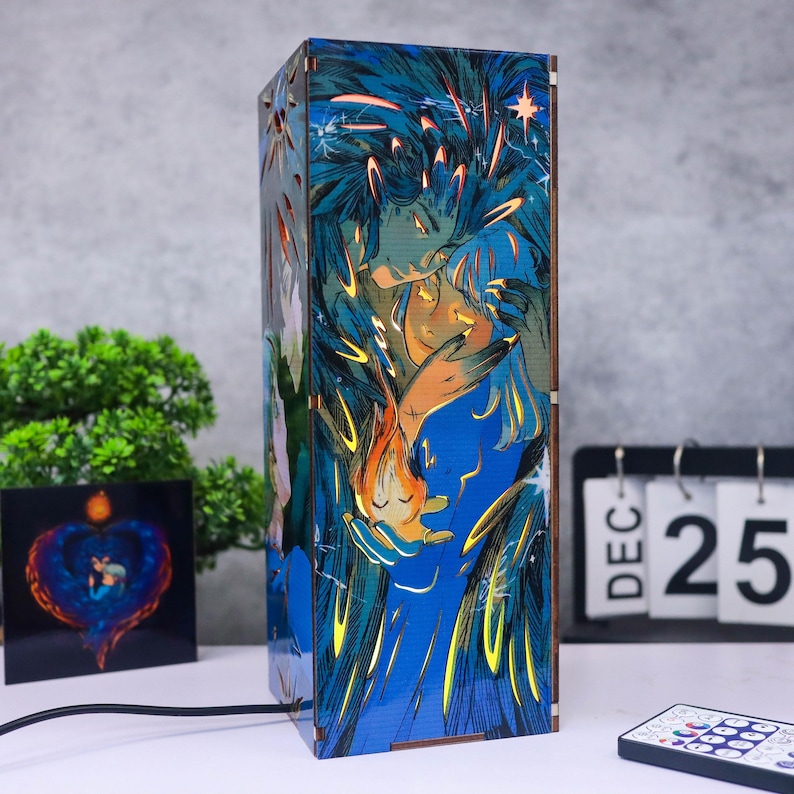 May include: A rectangular wooden lamp with a blue and orange illustration of two figures embracing. The lamp has a warm glow and is plugged in with a black cord. A small square art piece and a calendar are in the background.