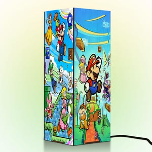 Puede incluir: L&aacute;mpara rectangular con ilustraciones coloridas de personajes de Super Mario. La l&aacute;mpara presenta a Mario, la Princesa Peach y otros personajes sobre un fondo azul. Un cable de alimentaci&oacute;n negro es visible en la base. La l&aacute;mpara mide unos 30 cm de alto.