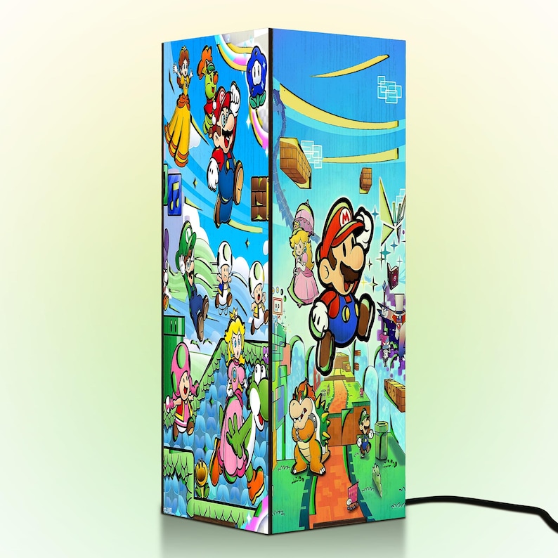 Puede incluir: L&aacute;mpara rectangular con ilustraciones coloridas de personajes de Super Mario. La l&aacute;mpara presenta a Mario, la Princesa Peach y otros personajes sobre un fondo azul. Un cable de alimentaci&oacute;n negro es visible en la base. La l&aacute;mpara mide unos 30 cm de alto.
