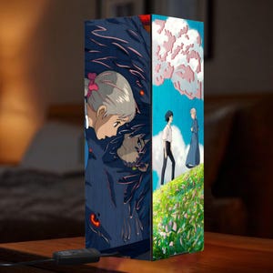 May include: A rectangular wooden lamp featuring anime artwork. One side depicts a dark, shadowy figure with a woman's face, while the other shows two characters on a grassy hill under a blue sky with clouds. The lamp has a black power cord.