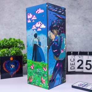 May include: A wooden lamp featuring anime-style artwork. The lamp's panels depict a couple embracing and a couple standing in a field with pink butterflies. The background is a blue sky with white clouds. A remote control and a calendar showing December 25 are in the foreground.
