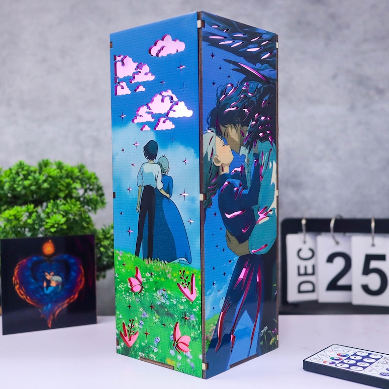 May include: A wooden lamp featuring anime-style artwork. The lamp's panels depict a couple embracing and a couple standing in a field with pink butterflies. The background is a blue sky with white clouds. A remote control and a calendar showing December 25 are in the foreground.