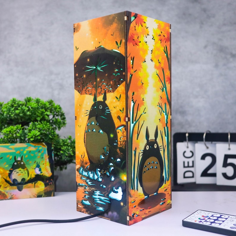 Puede incluir: L&aacute;mpara de madera con una colorida ilustraci&oacute;n de Totoro en un bosque. La l&aacute;mpara rectangular presenta dise&ntilde;os recortados que emiten luz. La paleta de colores incluye naranja, marr&oacute;n y verde.