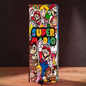 Puede incluir: Una caja rectangular cubierta de pegatinas coloridas con personajes de Super Mario. El texto "SUPER MARIO" se muestra de forma destacada en una fuente vibrante y multicolor. La caja est&aacute; sobre una superficie de madera con un fondo oscuro.