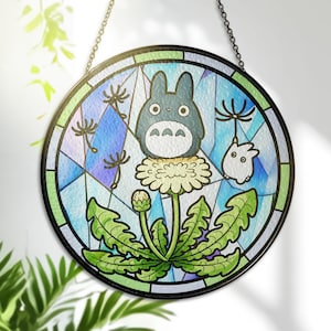 May include: Round stained glass panel featuring a cartoon Totoro character on a dandelion. The design includes a blue and green background with a black border and chain for hanging. The artwork is a colorful, whimsical design.