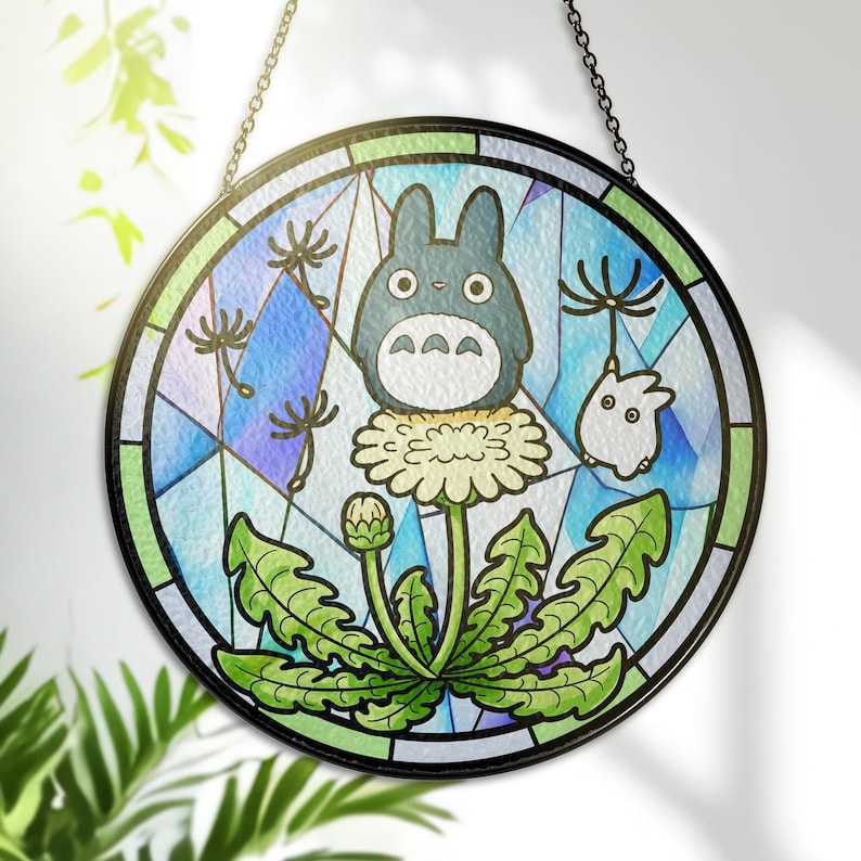 May include: Round stained glass panel featuring a cartoon Totoro character on a dandelion. The design includes a blue and green background with a black border and chain for hanging. The artwork is a colorful, whimsical design.