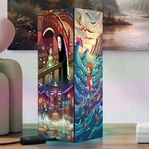 May include: A rectangular lamp featuring vibrant anime-style illustrations on its sides. One side depicts a character with a white mask and a girl on a wooden bridge, while another side shows a small character surfing on blue ocean waves with fish.