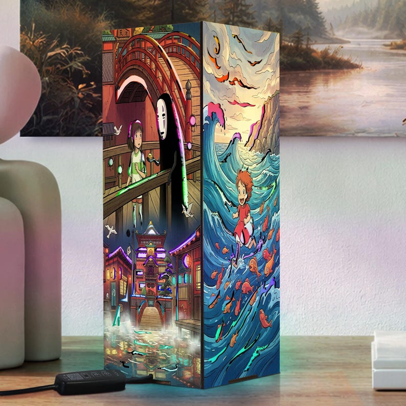 May include: A rectangular lamp featuring vibrant anime-style illustrations on its sides. One side depicts a character with a white mask and a girl on a wooden bridge, while another side shows a small character surfing on blue ocean waves with fish.