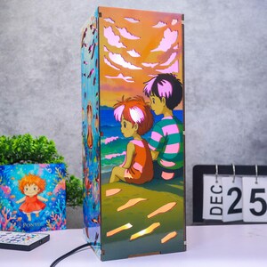 May include: A rectangular wooden lamp featuring a colorful illustration of two animated characters sitting by the ocean under a sunset sky. The side panel displays a character in a red dress underwater. The lamp emits a warm glow through cutouts.