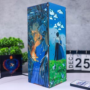 May include: A rectangular wooden lamp featuring anime-style artwork. The lamp's panels depict a couple, a blue sky with clouds, and butterflies. The artwork is primarily blue, orange, and green. A remote control and a calendar showing December 25 are in the foreground.