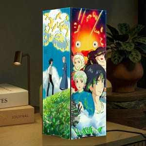 May include: A rectangular lamp featuring colorful anime artwork. One side depicts a scene with characters on a grassy hill under a blue sky. The other side showcases a fiery character and other anime figures. The lamp is on a wooden surface.