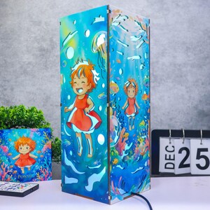 May include: A rectangular lamp featuring a vibrant underwater scene with a cartoon character in a red dress swimming among colorful fish, coral, and bubbles. The design includes cut-out shapes and a matching square card with the text PONYO.