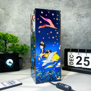 May include: A rectangular, illuminated wooden lamp featuring a night sky design with a flying witch and a black cat. The lamp has a blue background with stars and a flying bird. A remote control and a calendar showing December 25 are also visible.