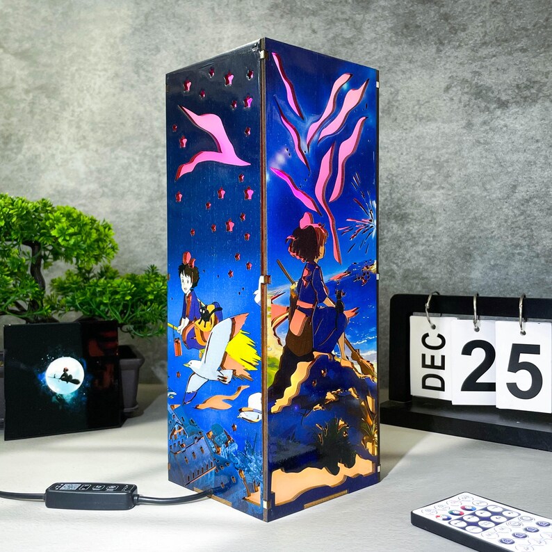 May include: A rectangular lamp with a blue and pink anime-style design. The lamp features cut-out shapes and illustrations of characters, birds, and fireworks. A remote control and a calendar showing December 25 are in the foreground.
