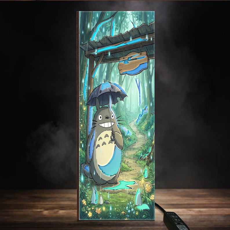 May include: A rectangular light panel featuring a colorful illustration of a grey, round creature with a white belly holding a dark umbrella in a rainy forest. The scene includes a wooden sign, green trees, small white spirits, and yellow flowers.