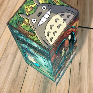 May include: A square wooden lamp featuring colorful illustrations of animated characters. The top panel displays a large grey creature with a wide smile and an acorn on its head. The side panels depict a forest scene and a bridge with a dark figure.