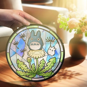 May include: Round, black clutch with a stained-glass design featuring a blue Totoro character, a white creature, and a dandelion. The design incorporates blue, green, and yellow colors. The clutch is held by a hand.
