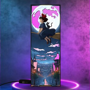 May include: A rectangular lamp featuring an anime-style illustration. A young witch with a red bow flies on a broom with a black cat against a pink moon. Below, a city street scene with pink-lit buildings and a reflective road.