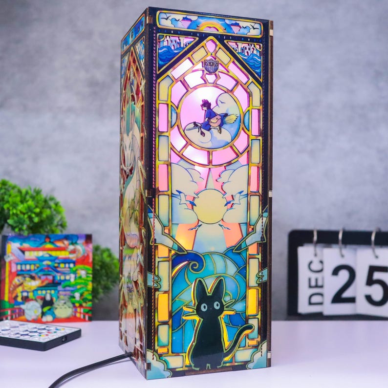 May include: A rectangular wooden lamp featuring a stained-glass style design with colorful illustrations of a black cat, a figure on a broomstick, and a sun. The lamp emits a warm glow, showcasing intricate cutouts and layered panels on its sides.