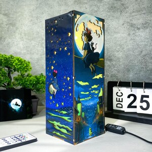May include: A rectangular wooden lamp featuring a painted anime scene with a witch flying on a broom in front of a full moon. The lamp has a starry night sky design on one side and a cityscape on another. The lamp is lit with small lights.