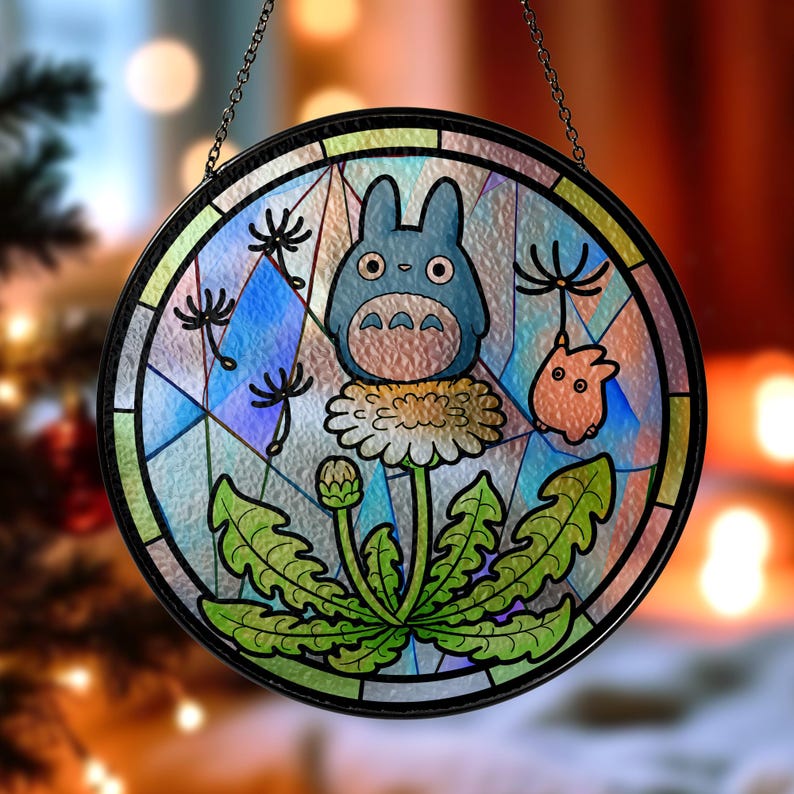 May include: Round stained glass window featuring a blue Totoro character and a small orange creature, set against a dandelion. The design is framed in black and hangs from a chain. The glass has a colorful, mosaic-like appearance.