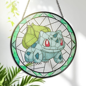 Puede incluir: Una vidriera redonda con un diseño de Bulbasaur. La obra de arte muestra un Bulbasaur azul claro con ojos rojos y detalles verdes, sobre un fondo blanco. La ventana está enmarcada con un borde negro y un borde decorativo verde.