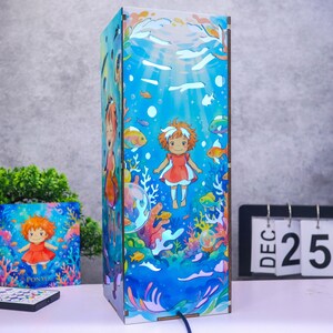 May include: A rectangular wooden lamp featuring a vibrant underwater scene with a cartoon character in a red dress surrounded by colorful coral, various fish, and bubbles. The design includes light blue water tones and a dark brown frame structure.