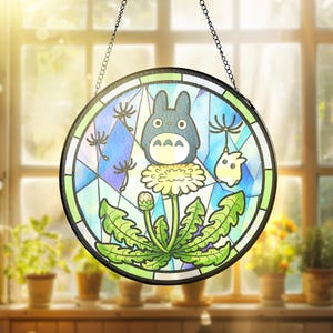 May include: Round stained glass panel featuring a cartoon character with a dandelion design. The artwork includes a blue and green color scheme, with a black chain for hanging. The panel is displayed in front of a window.