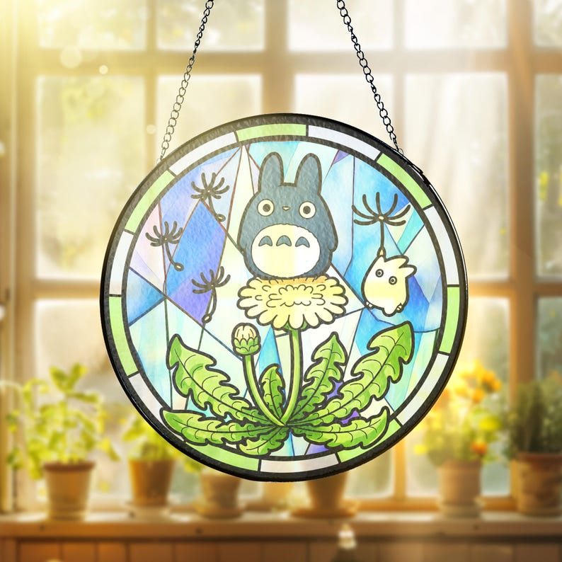 May include: Round stained glass panel featuring a cartoon character with a dandelion design. The artwork includes a blue and green color scheme, with a black chain for hanging. The panel is displayed in front of a window.
