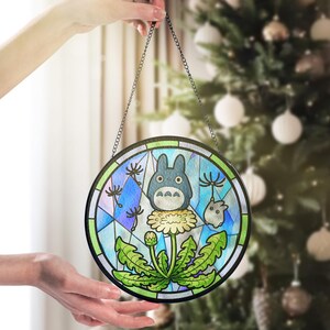 May include: Round stained glass panel featuring a blue Totoro character on a dandelion. The artwork has a black border and is suspended by a chain. The background includes a geometric pattern with blue, green, and white colors.