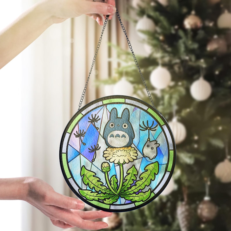 May include: Round stained glass panel featuring a blue Totoro character on a dandelion. The artwork has a black border and is suspended by a chain. The background includes a geometric pattern with blue, green, and white colors.