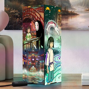 Puede incluir: Una l&aacute;mpara rectangular con ilustraciones de anime coloridas. La l&aacute;mpara muestra escenas y personajes de una popular pel&iacute;cula de animaci&oacute;n, con ilustraciones vibrantes de un puente, edificios y un drag&oacute;n. La l&aacute;mpara tiene un cable de alimentaci&oacute;n negro.