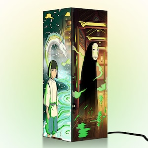 Puede incluir: L&aacute;mpara rectangular con ilustraciones de estilo anime. Un lado representa a un joven y un drag&oacute;n blanco cerca de un r&iacute;o. El otro lado muestra una figura con capa y m&aacute;scara negras. La l&aacute;mpara tiene un cable de alimentaci&oacute;n negro.