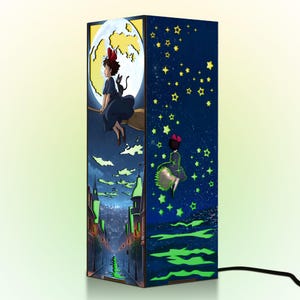 May include: A rectangular lamp featuring illustrations from an animated film. One side depicts a girl on a broom in front of a moon, and another shows her flying among stars. The lamp has a black power cord.
