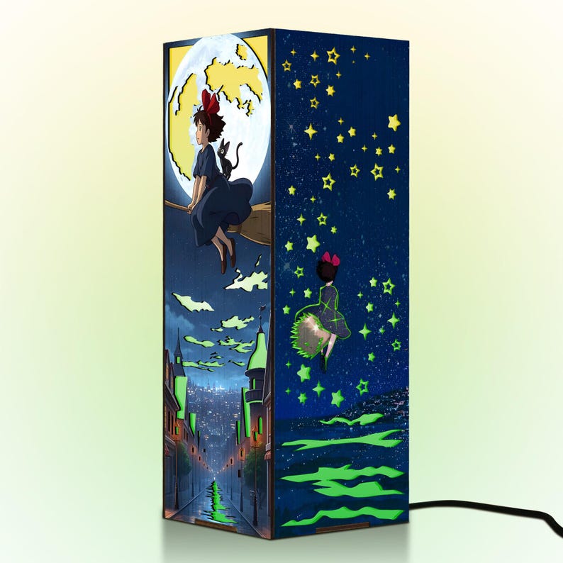 May include: A rectangular lamp featuring illustrations from an animated film. One side depicts a girl on a broom in front of a moon, and another shows her flying among stars. The lamp has a black power cord.