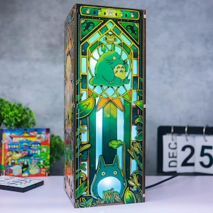 May include: A rectangular wooden lamp featuring a stained-glass style design with vibrant green and yellow hues. The front panel displays a large green creature with pointed ears and a white belly, surrounded by intricate botanical patterns and foliage.
