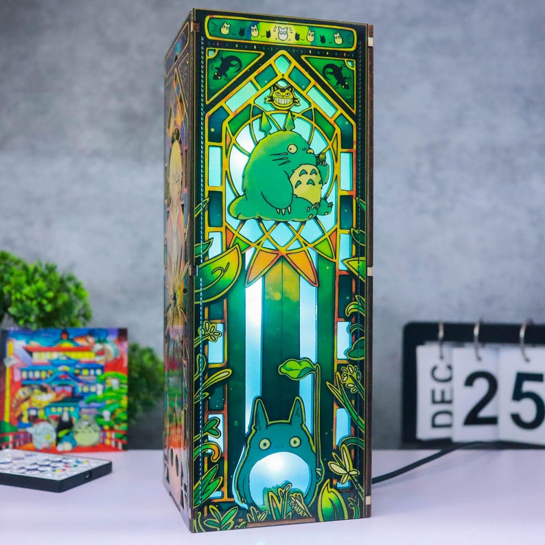 May include: A rectangular wooden lamp featuring a stained-glass style design with vibrant green and yellow hues. The front panel displays a large green creature with pointed ears and a white belly, surrounded by intricate botanical patterns and foliage.