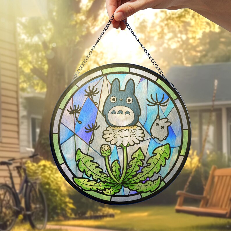 May include: Round stained glass panel featuring a blue Totoro character with a white flower and green leaves. The panel has a black metal frame and a chain for hanging. The background is a colorful mix of blue, green, and white.