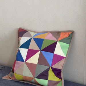 Könnte beinhalten: Quadratisches Zierkissen mit geometrischem Patchwork-Design. Das Kissen weist eine Vielzahl von Farben auf, darunter Blau, Rosa, Grün und Orange, die in einem dreieckigen Muster angeordnet sind. Es hat einen braunen Rand und liegt auf einer gemusterten Oberfläche.