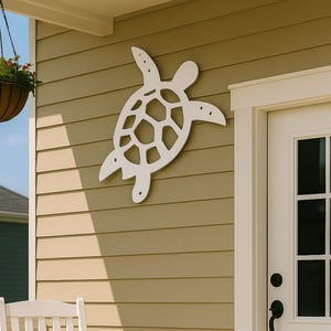 May include: A white wooden rocking chair sits on a brown porch, next to a white turtle-shaped wall decoration. A hanging basket with flowers and a white door with black hardware are also visible. The house has tan siding.