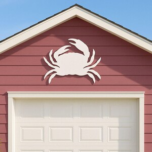 May include: A white crab-shaped wall decoration mounted on a red-painted exterior wall above a white garage door. The crab has detailed claws and legs. The house trim is white, and the sky is blue.