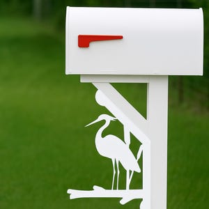 May include: White mailbox with a red flag, mounted on a white post with a decorative heron silhouette. The mailbox is set against a backdrop of green grass and a gravel driveway.