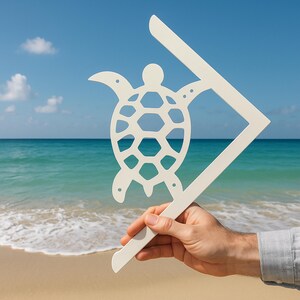 May include: White metal turtle cutout attached to a white metal corner bracket. The bracket is held in a hand against a background of a beach and ocean.