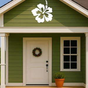 Puede incluir: Una decoraci&oacute;n de flor de hibisco blanca est&aacute; montada en el revestimiento verde de una casa. La casa tiene una puerta blanca con una corona, una ventana y una peque&ntilde;a planta en maceta. El porche tiene escalones de madera.