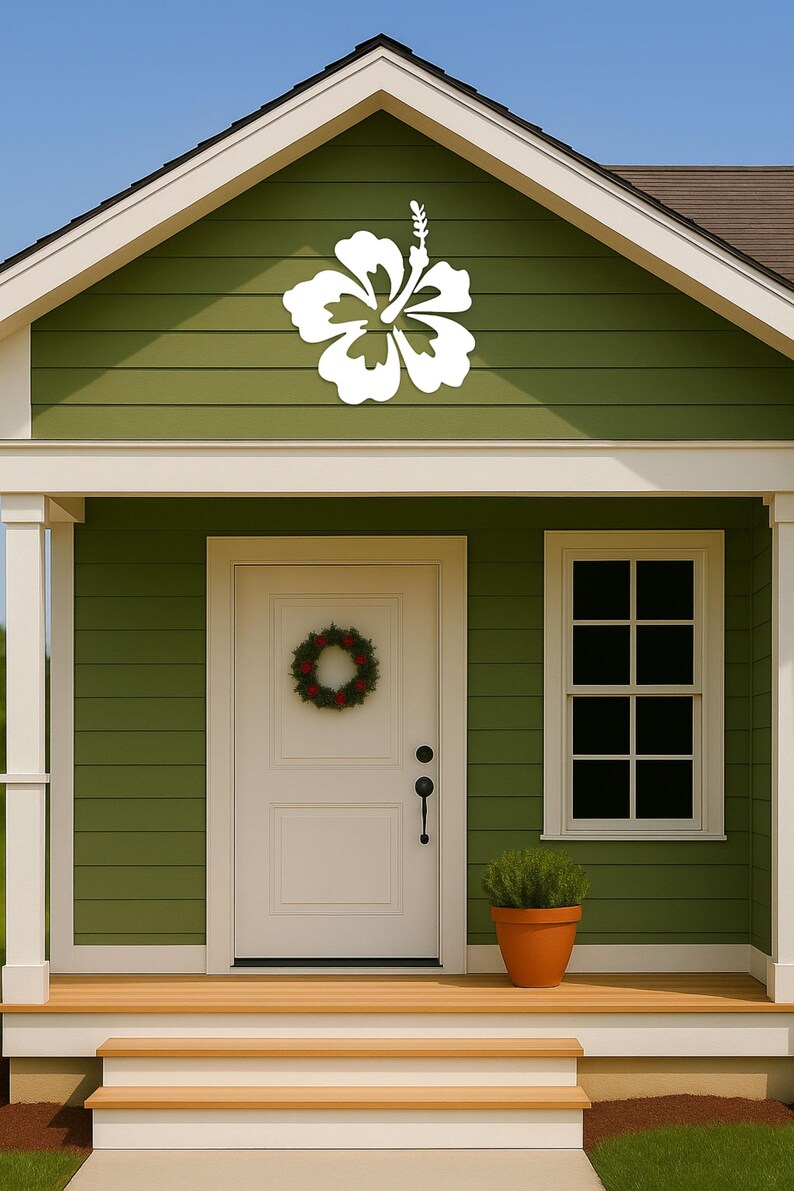 Puede incluir: Una decoraci&oacute;n de flor de hibisco blanca est&aacute; montada en el revestimiento verde de una casa. La casa tiene una puerta blanca con una corona, una ventana y una peque&ntilde;a planta en maceta. El porche tiene escalones de madera.