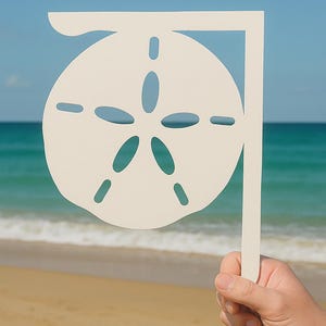 May include: A cream-colored sand dollar cutout held against a backdrop of a beach and ocean. The sand dollar has a star-like pattern of holes and is attached to a vertical handle. The ocean is turquoise, and the sand is tan.