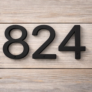 May include: Black metal house numbers 824 mounted on a light wood background. The numbers are in a modern font and have a matte finish. The wood grain is visible.