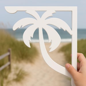 May include: White wooden shelf bracket with a palm tree cutout design. The bracket is held in a hand against a blurred background of a beach.
